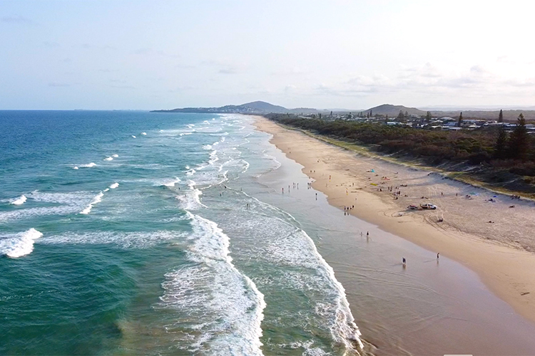 An aerial drone view looking down a long, sandy beach coastline with ocean waves rolling ashore, scattered beachgoers, and a coastal town stretching into the distance.