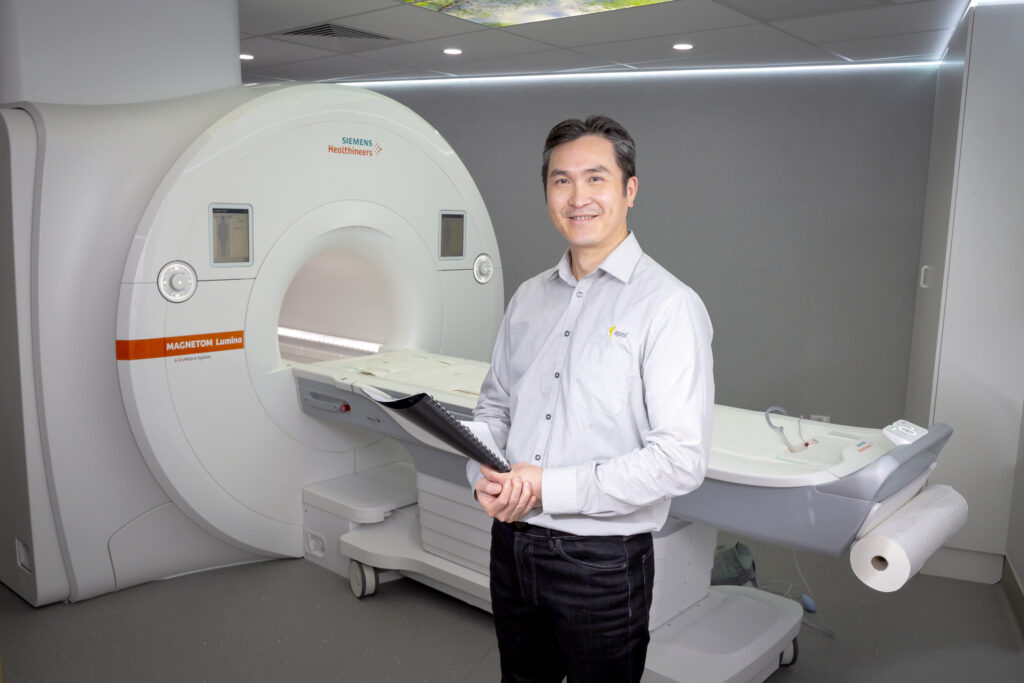 A healthcare professional standing beside an MRI scanner in a medical imaging suite, holding patient documentation.