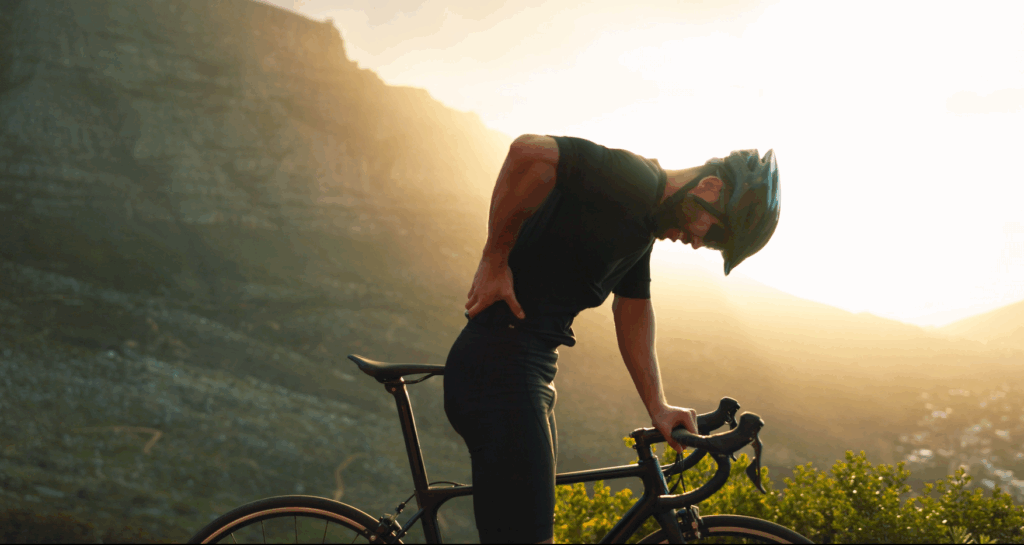 Biker holding his bike with one hand and his back with the other, showing he is in pain. Sunset and mountains in the background.
