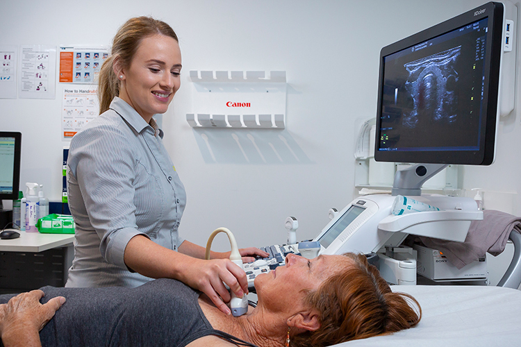 A sonographer performs an ultrasound examination on a patient’s neck using a handheld transducer while monitoring the scan on an ultrasound machine.