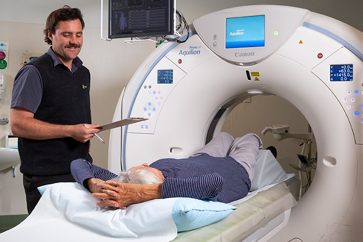 A patient lies on a CT scanner table while a radiographer stands nearby holding a clipboard.