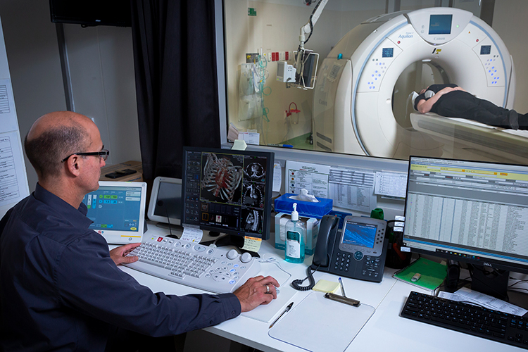 A radiographer operates a control workstation while a patient lies in a CT scanner on the other side of a viewing window.