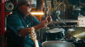 A musician with a drum kit in a workshop filled with tools and equipment, holding drumsticks during a rehearsal or performance setup.