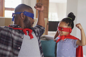 An adult and a child wearing superhero capes and eye masks, raising their arms while playing together at home.