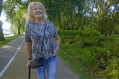 An older adult walking along a paved path in a park, using a walking cane for support.