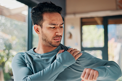 A person indoors touching their shoulder while examining arm movement, representing shoulder discomfort or joint pain.